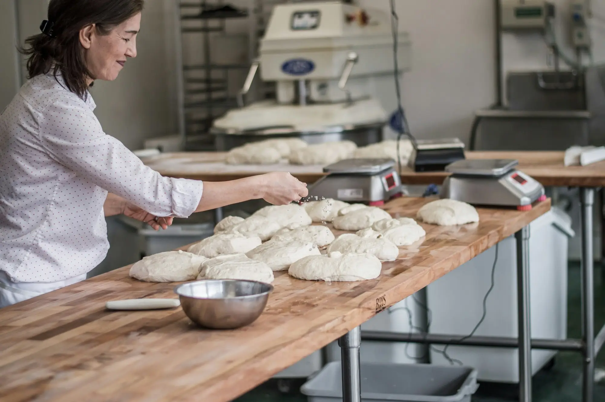 Bakery owner showing a new employee how to shape dough at a floured bench